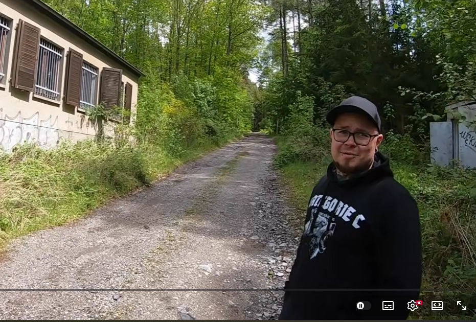 an einem "Waldweg/Pfad/Straße" steht ein Typ schwarzem Cap, Brille, ein Lippenpiercing (?) sowie Tätowierungen am Hals und Hoodie mit dem Label "Kategorie C" und schaut direkt in die Kamera.
Drum herum Wald und auf der rechten Seite ein herunter gekommenes Gebäude. an einem "Waldweg/Pfad/Straße" steht ein Typ schwarzem Cap, Brille, ein Lippenpiercing (?) sowie Tätowierungen am Hals und Hoodie mit dem Label "Kategorie C" und schaut direkt in die Kamera.
Drum herum Wald und auf der rechten Seite ein herunter gekommenes Gebäude.